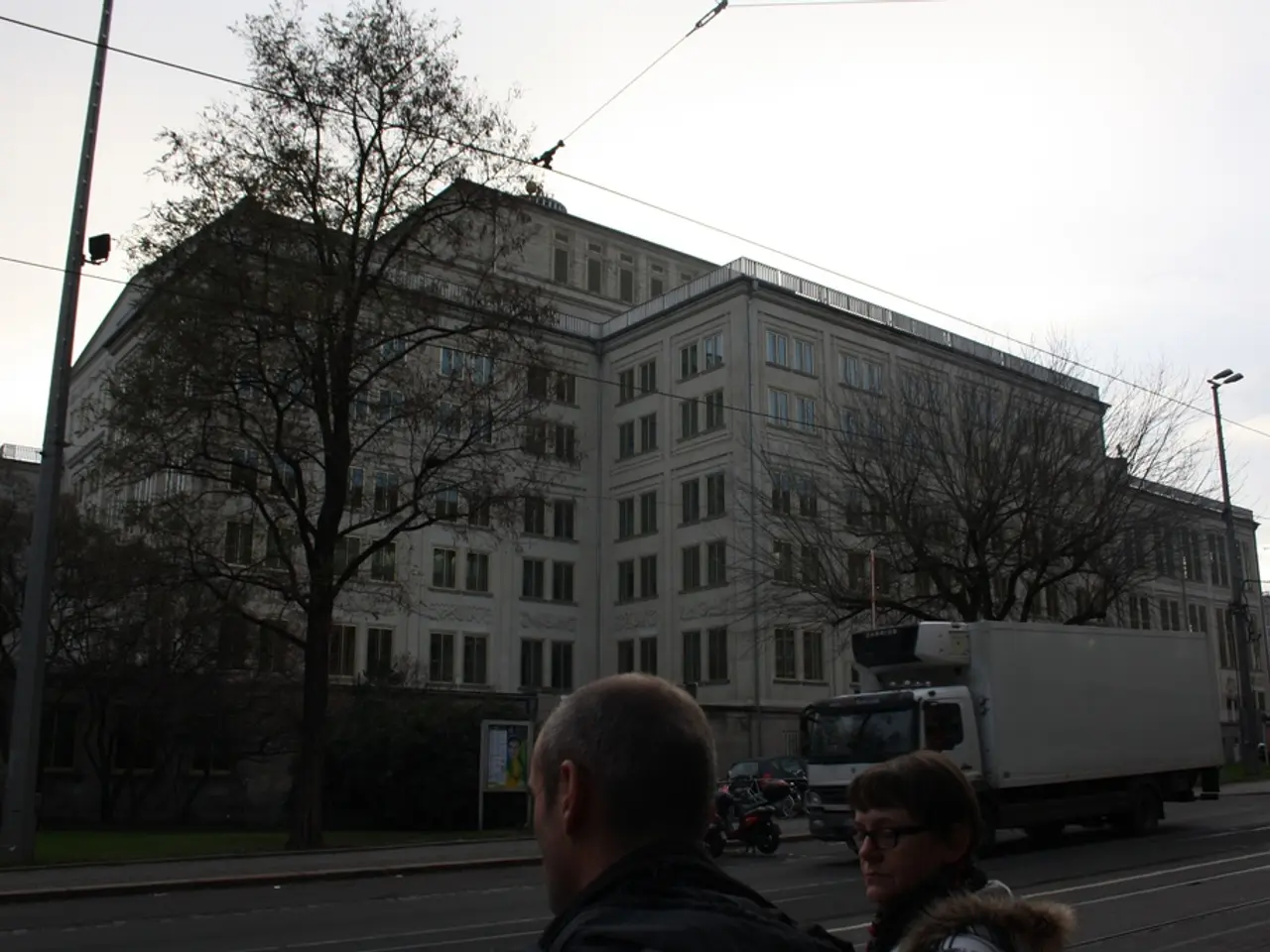 In this image we can see sky, electric poles, electric cables, buildings, trees, advertisement...