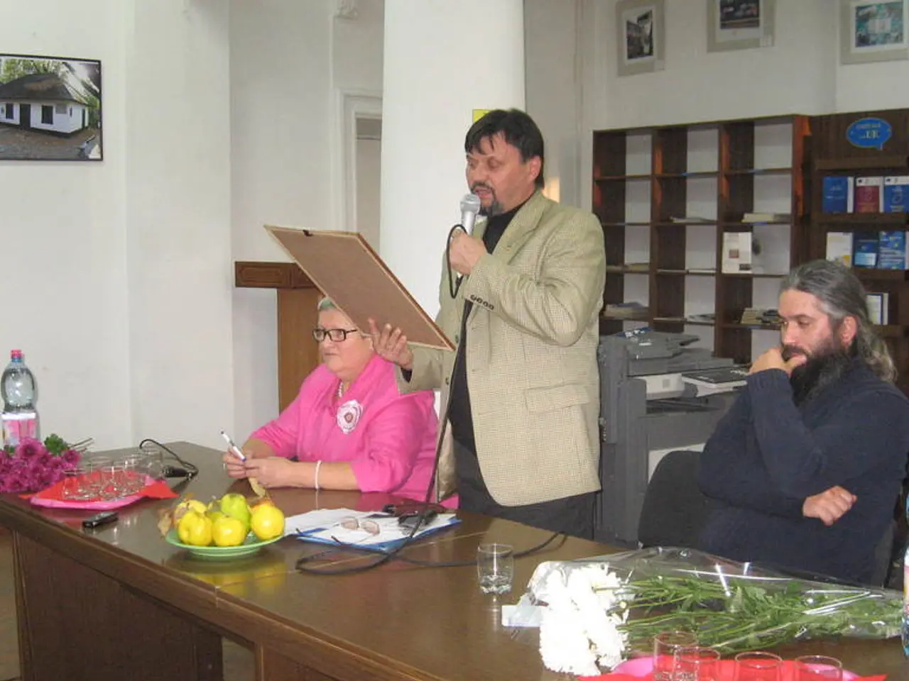 There are two persons sitting on the chairs,one person is standing,holding plank and mike in his...