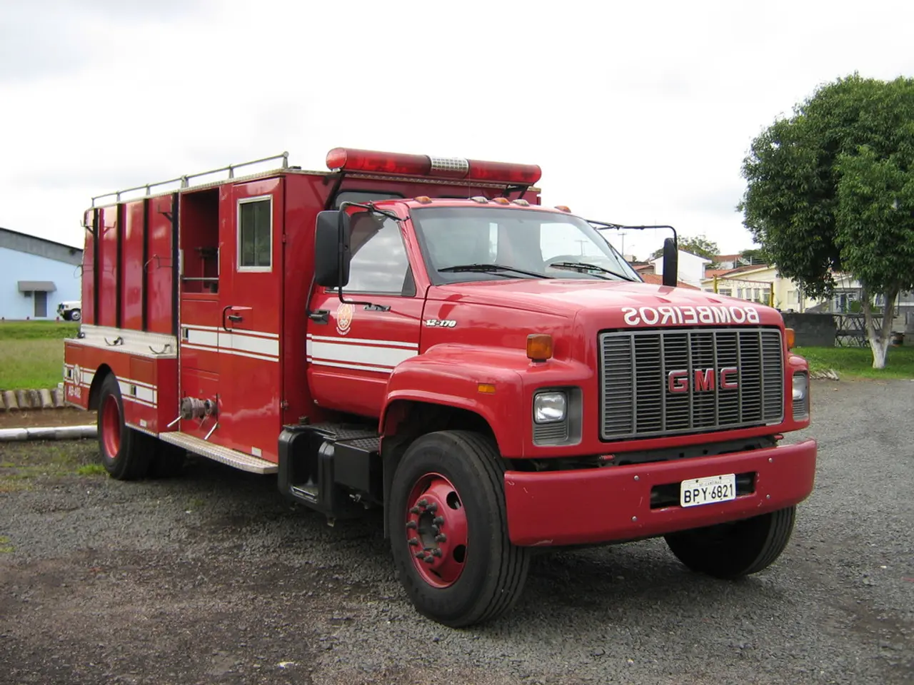 In this image we can see motor vehicles on the road, trees, grass, buildings and sky with clouds.