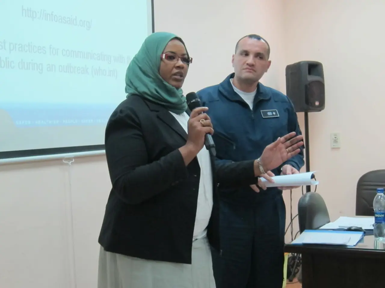 In this image a woman is speaking with the help of microphone, besides to her a man is standing,...