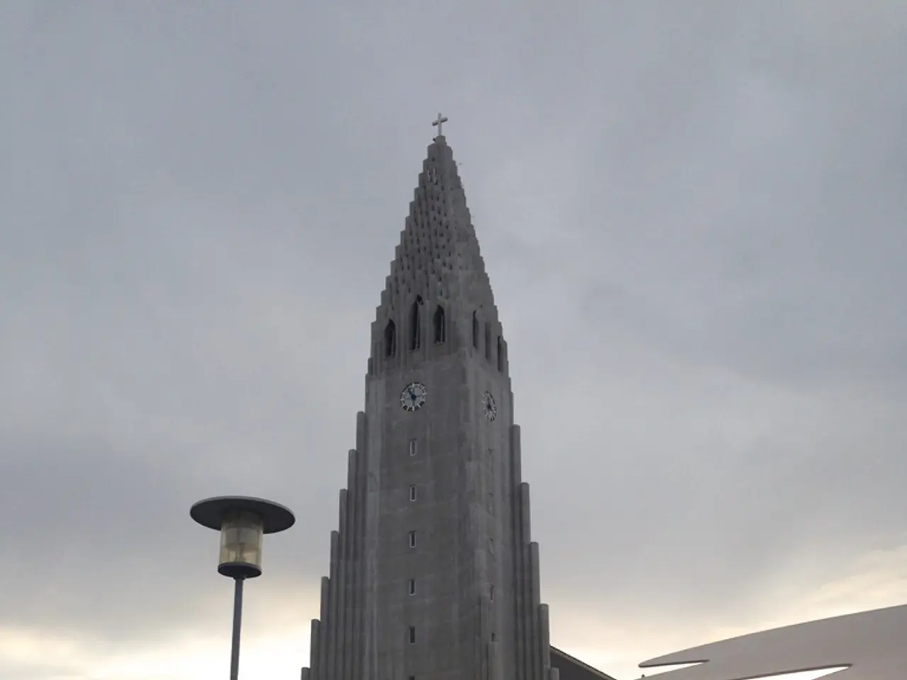 In this image there is a building with clock on it, also there are some trees and electrical pole...